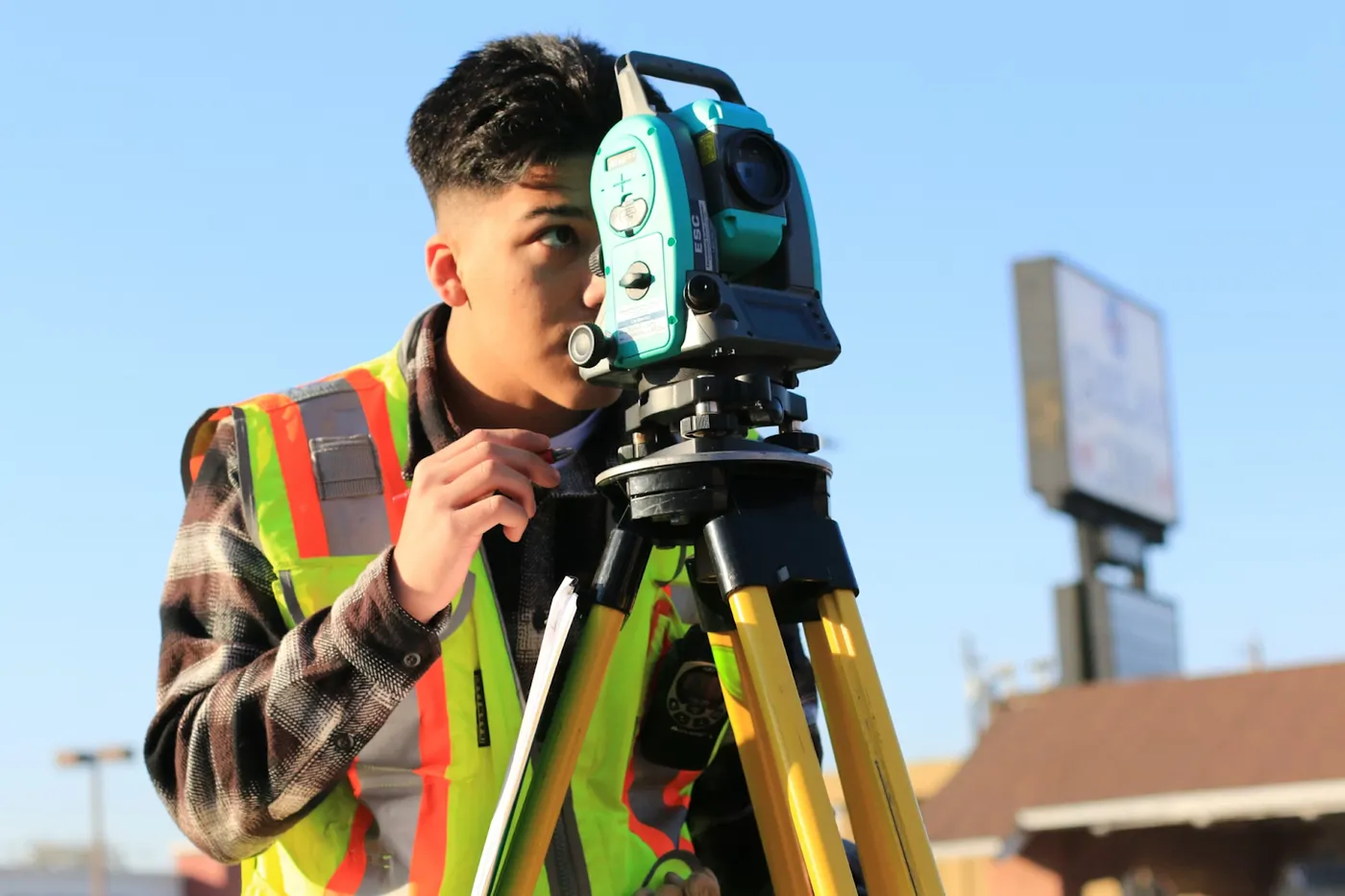 A photo of a surveyor in a high-visibility vest looking past a total station mounted on a yellow tripod