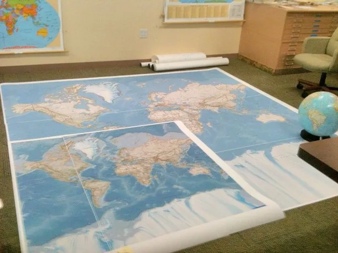 A photo of large printed world map tile sheets laid out on an office floor beside a desk globe